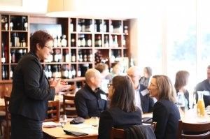 A woman is standing in front of a group of people sitting at tables in a restaurant.