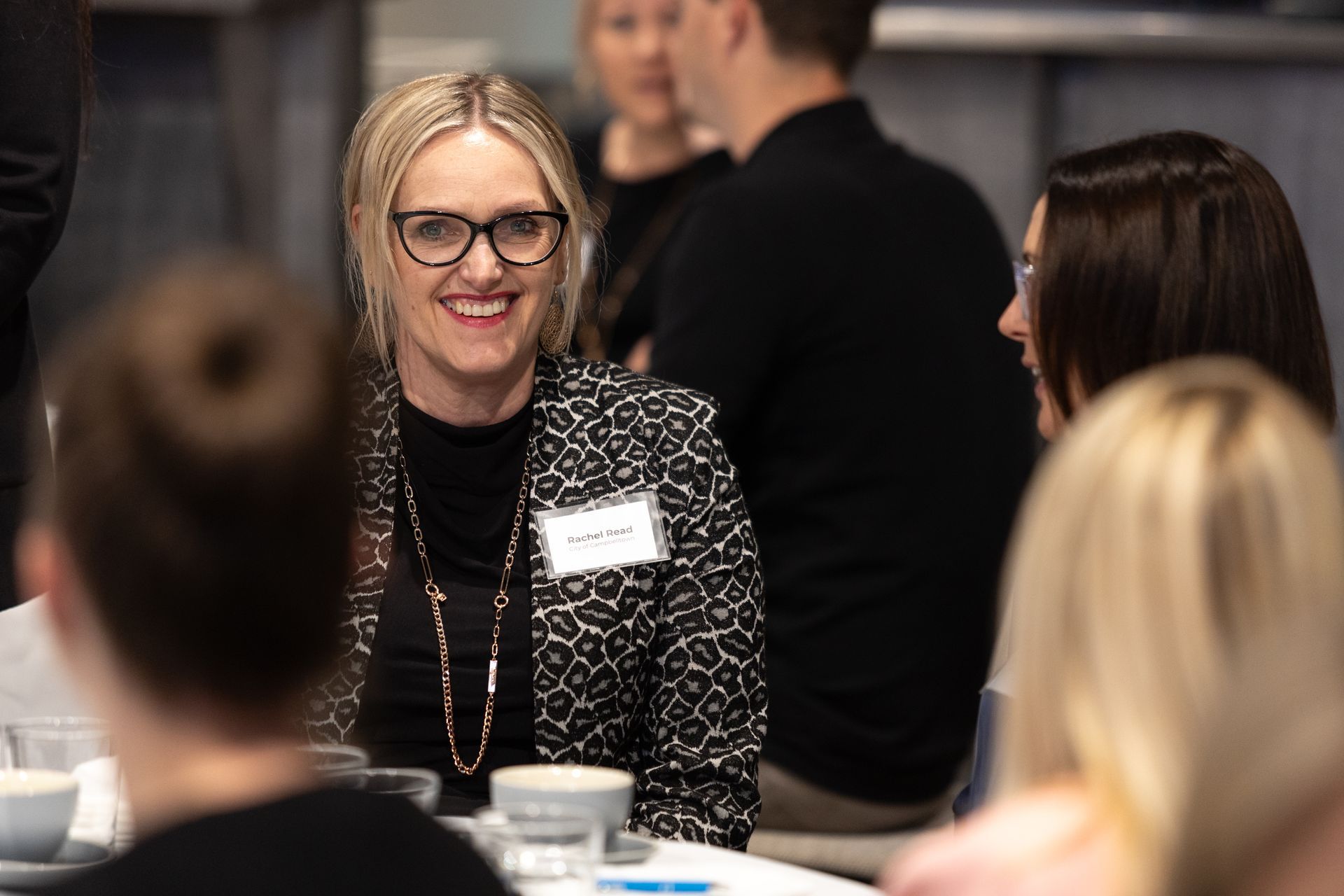 A woman wearing glasses is smiling while sitting at a table with other people.