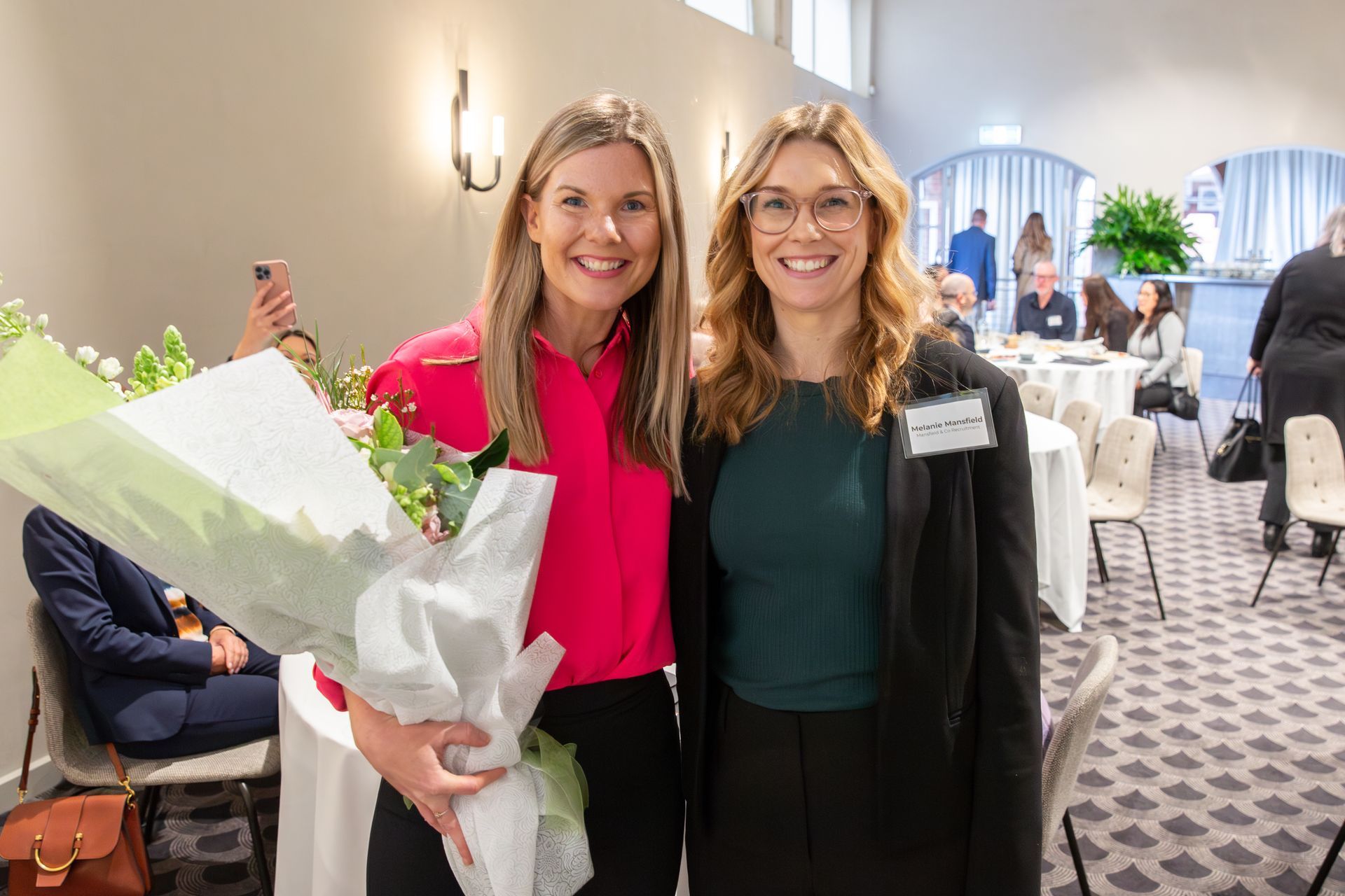 Two women are standing next to each other in a room holding a bouquet of flowers.