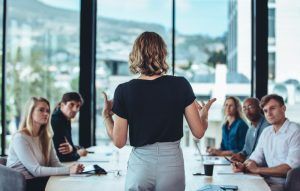 A woman is giving a presentation to a group of people sitting around a table.