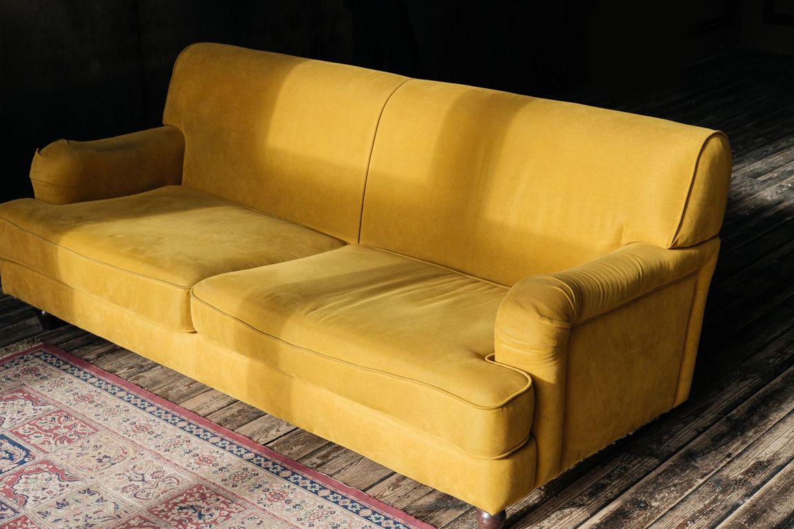 Un divano imbottito color giallo senape è posizionato su un pavimento in legno parzialmente coperto da un tappeto a motivi geometrici su uno sfondo scuro.