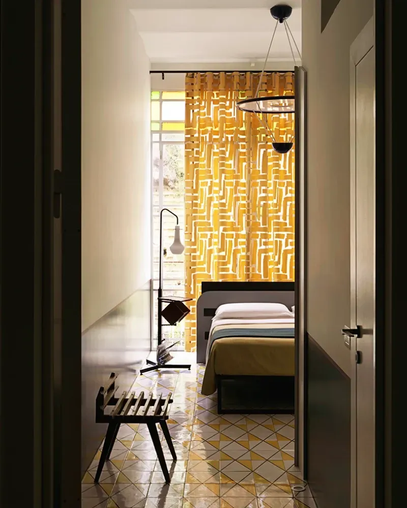 Una camera d'albergo vista dal corridoio, con un letto, tende gialle a motivi geometrici, una lampada, un piccolo sgabello e pavimento piastrellato.