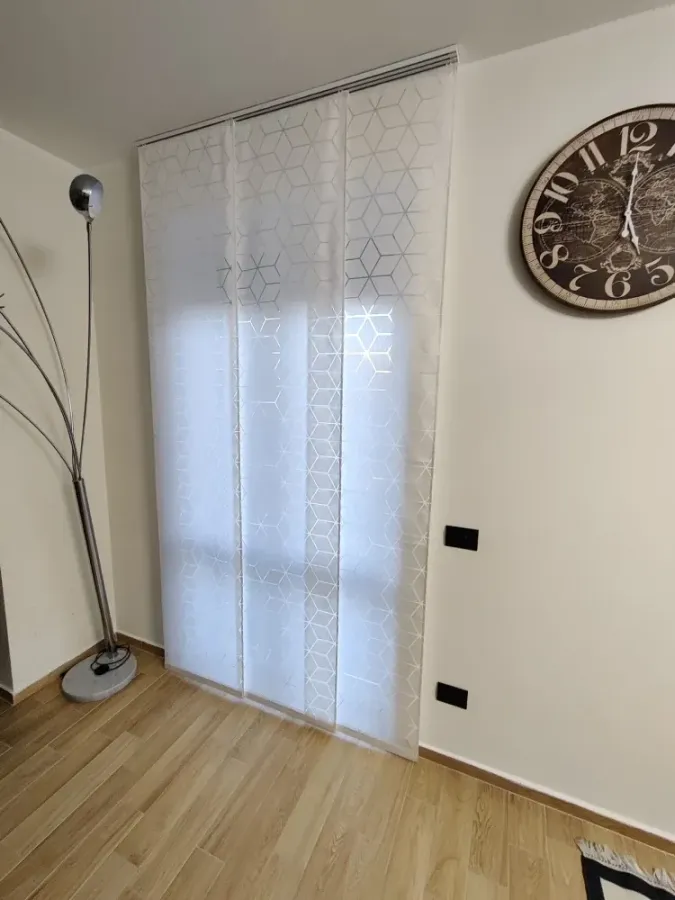 Una tenda a pannello bianca con motivo geometrico copre una finestra accanto a una moderna lampada da terra e a un grande orologio rotondo da parete.
