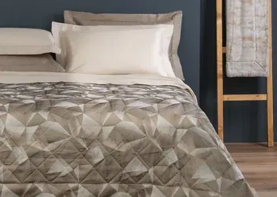 Un letto con un piumone a motivi geometrici grigi e bianchi, cuscini color crema e una coperta dalla trama particolare appesa a un supporto in legno.