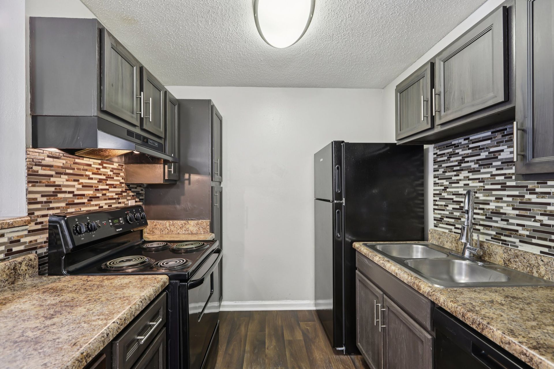 A galley kitchen with dark wood cabinets, granite countertops, black appliances, and a decorative tile backsplash.
