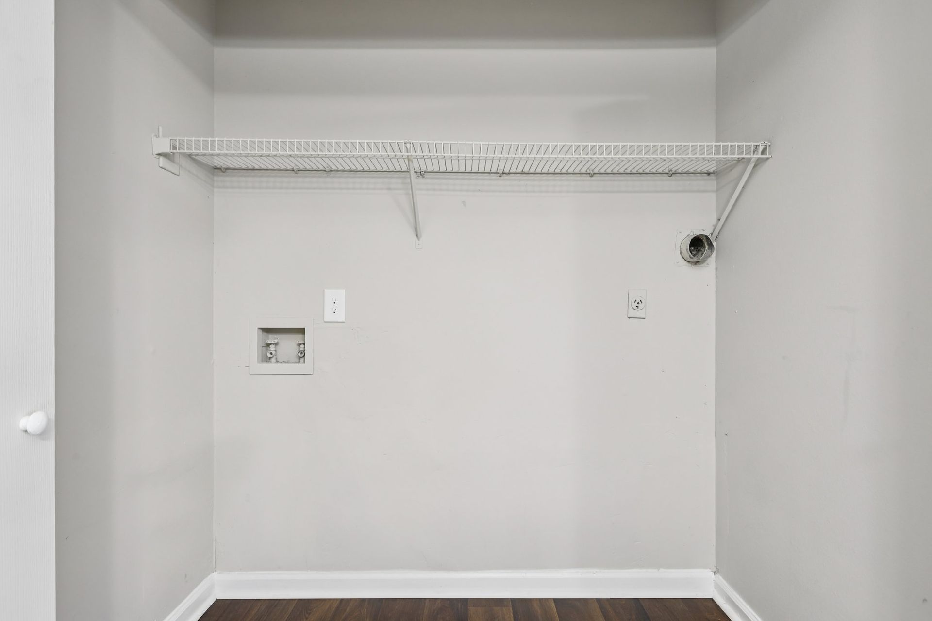 A laundry closet with a wire shelf, water hookups, and an exhaust vent against a plain white wall.