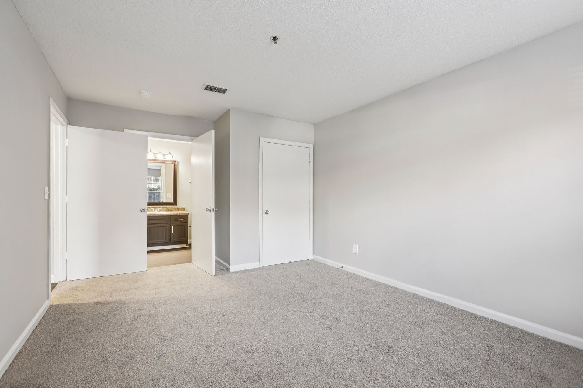 A neutral-toned empty bedroom with grey carpet, white walls, a closet door, and an open doorway leading to a bathroom.