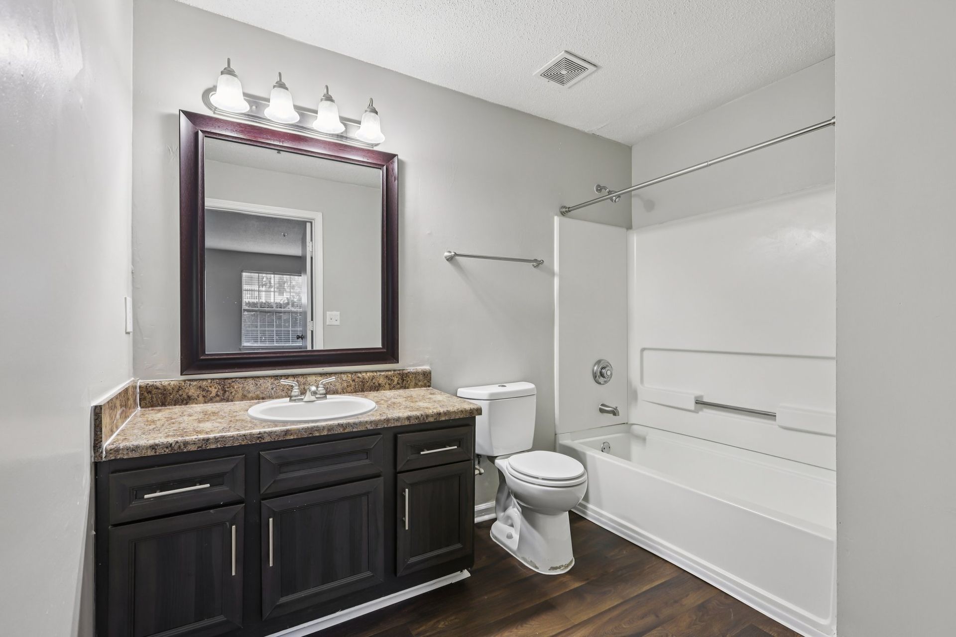 A modern bathroom with dark wood cabinets, granite countertops, a framed mirror, and a white tub-shower combo.