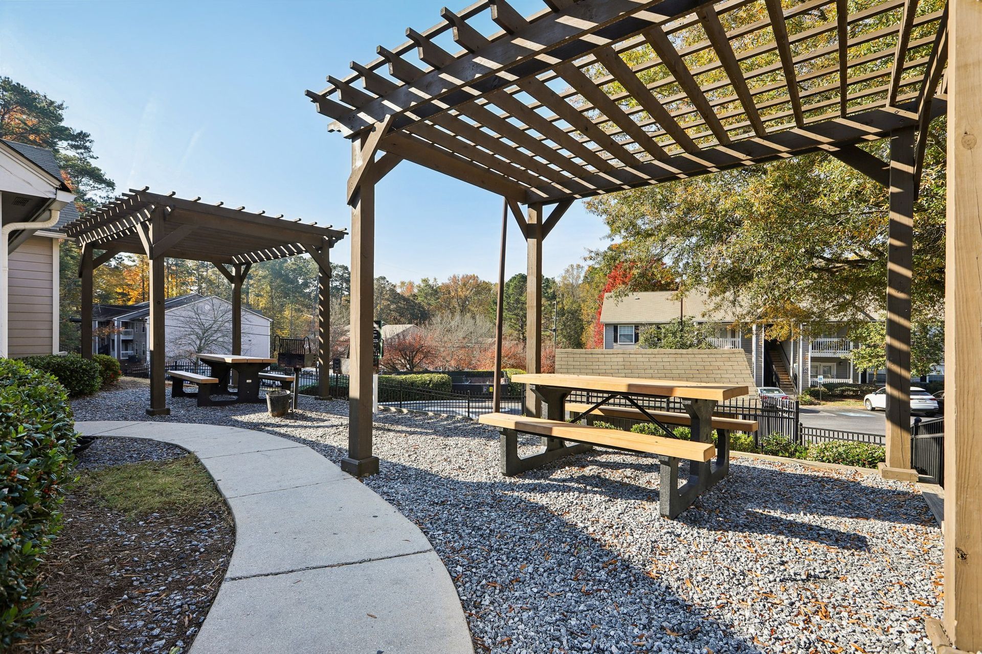 Two wooden pergolas with picnic tables stand over a gravel patio with a winding concrete path under a clear blue sky.
