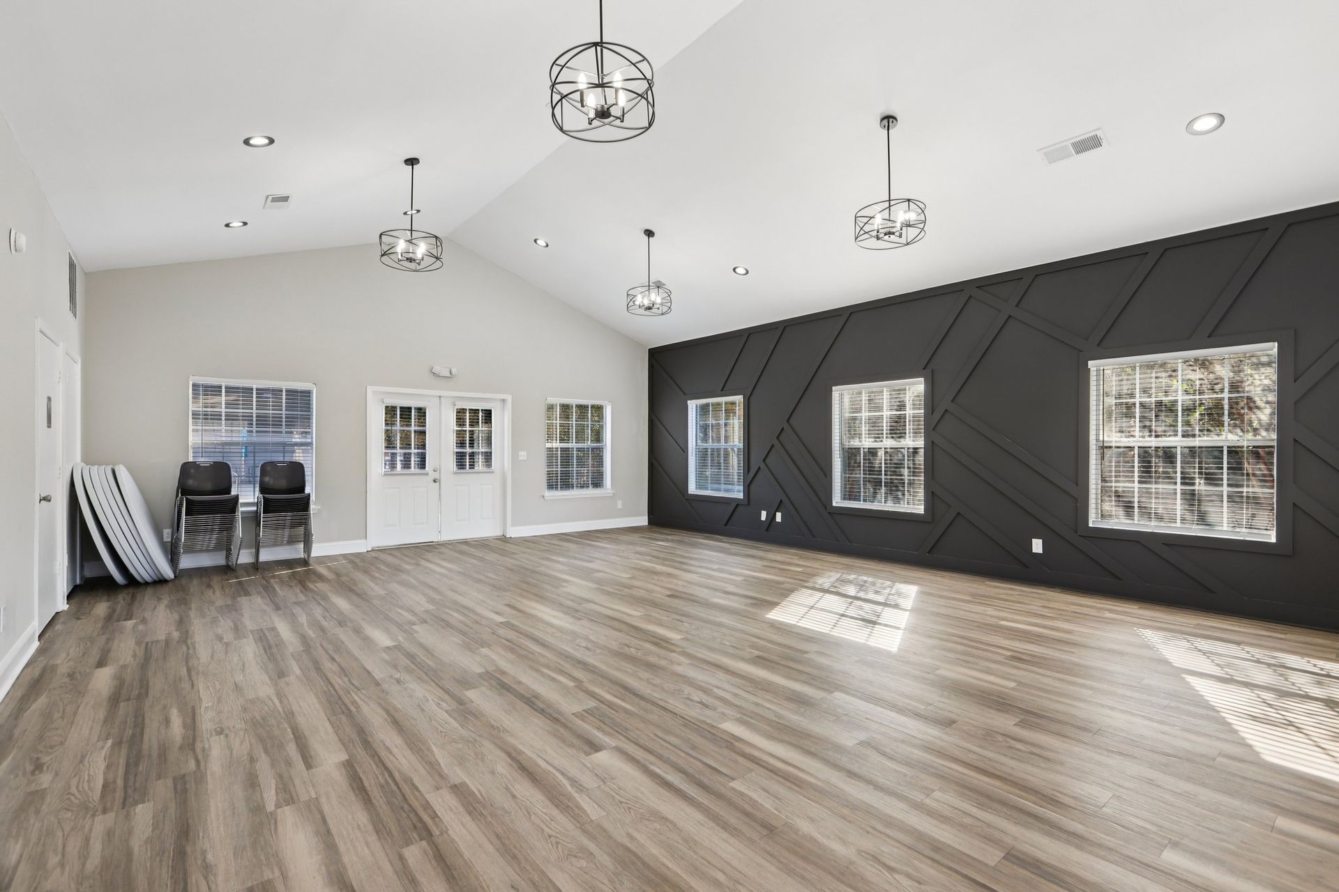 A bright, spacious community room with wood-look floors, a vaulted ceiling, four pendant lights, and a black accent wall.