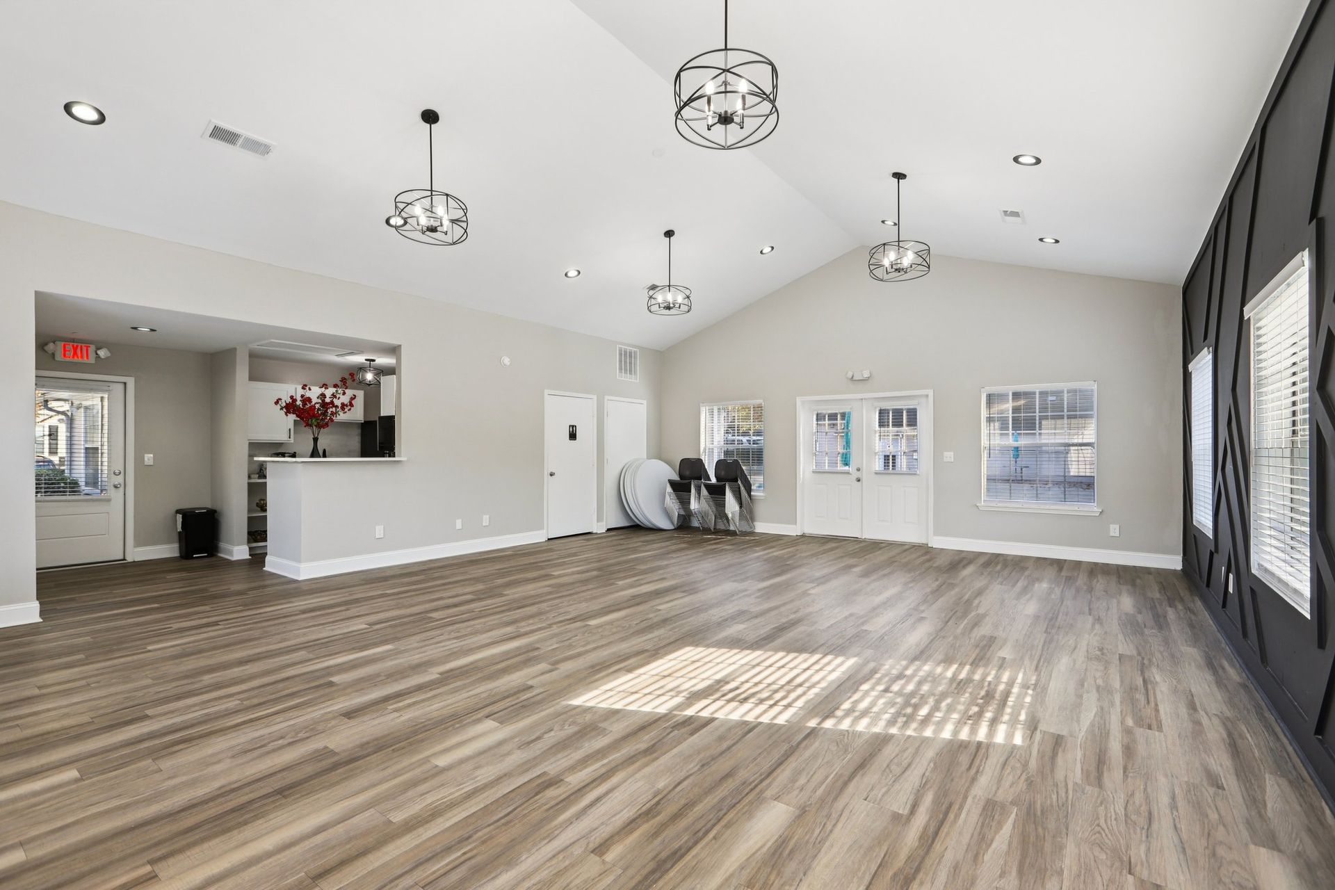 A spacious, bright community room with wood-look flooring, neutral walls, four pendant lights, and a small kitchen area.