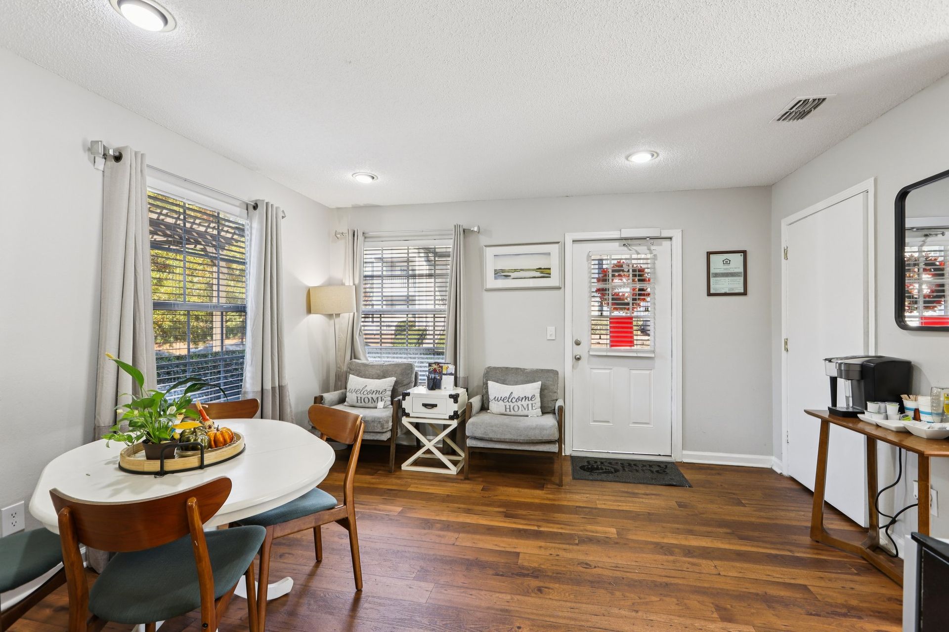 A bright dining area featuring a round white table, grey accent chairs, wood floors, and a white front door.