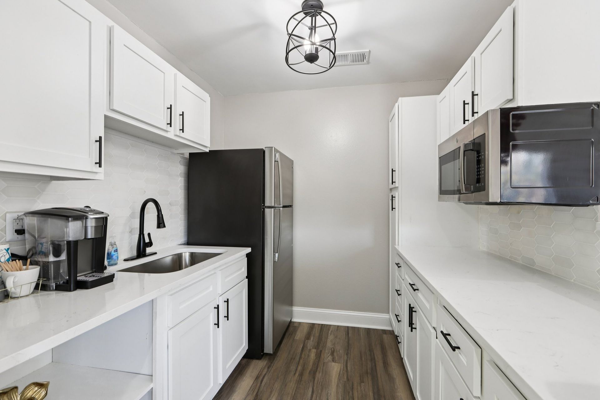 A galley kitchen with white cabinets, light countertops, black appliances, and a modern metal pendant light.
