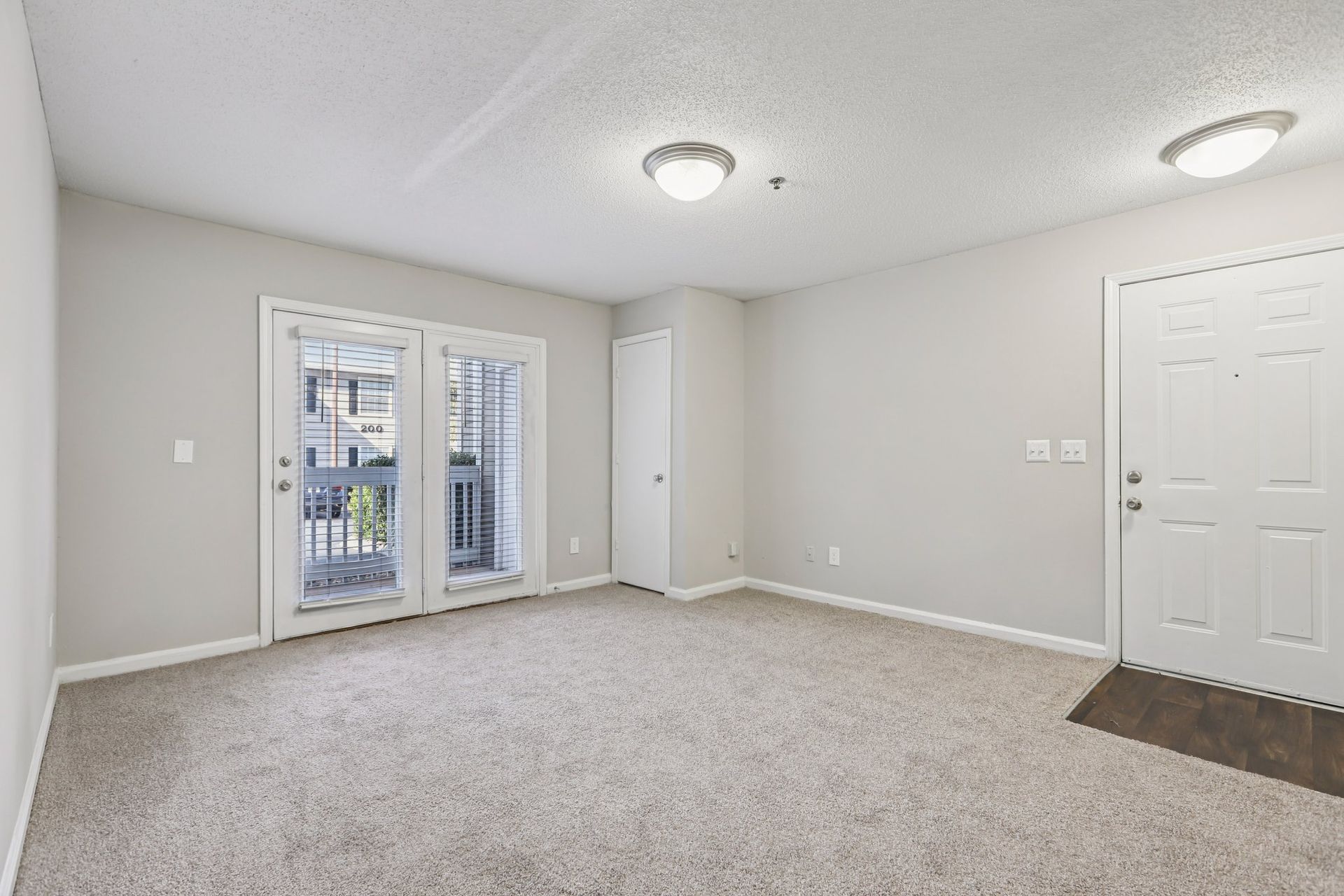 Empty room with light-colored carpet and walls, white ceiling lights, a white door, and sliding glass balcony doors.