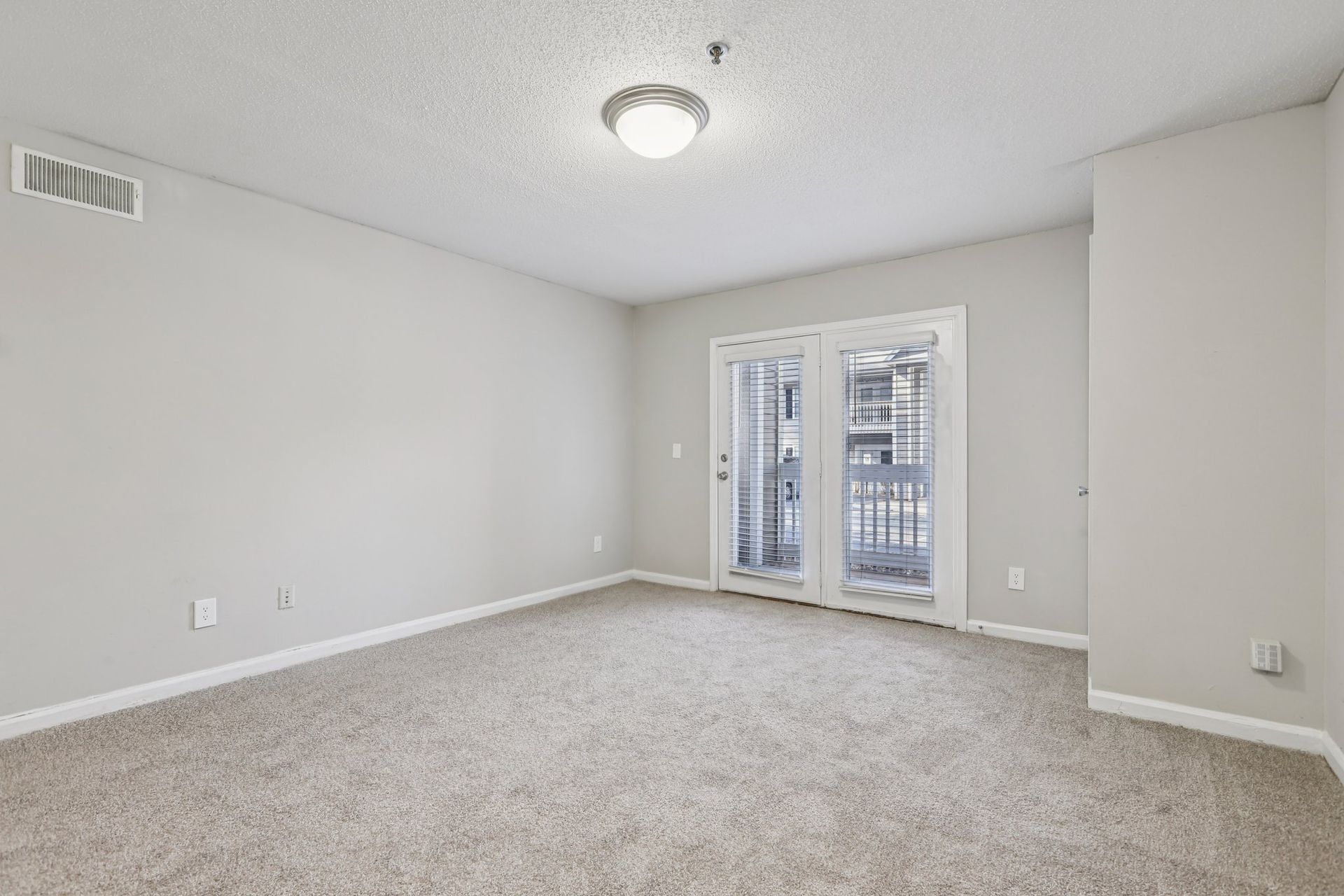 An empty, neutrally painted bedroom with beige carpet, a ceiling light, and glass patio doors leading to a balcony.