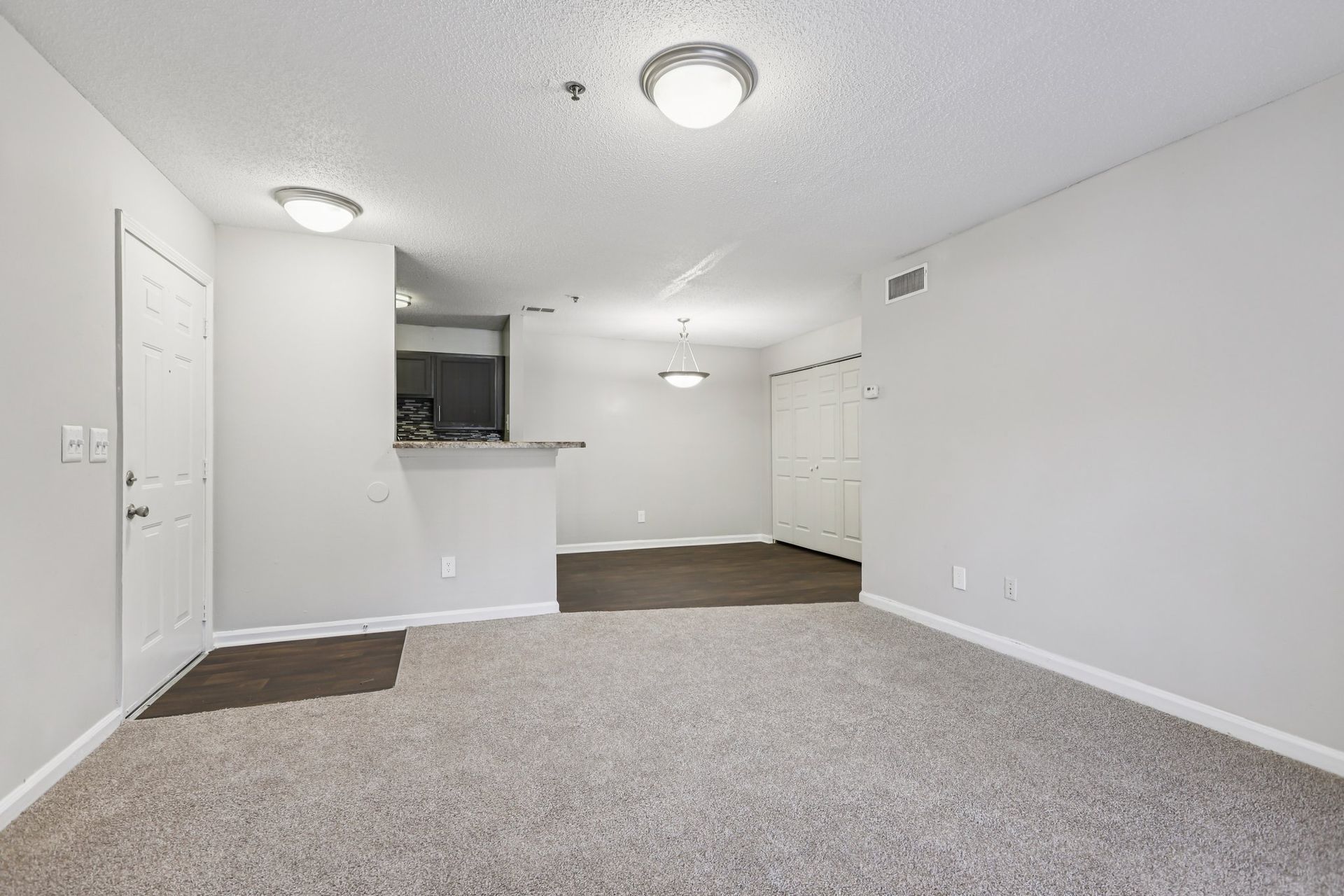 Empty living room with light gray walls, beige carpet, a doorway, and an open kitchen area in the background.
