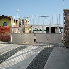 Cancello e recinzione in metallo bianco chiusi all'ingresso di un'abitazione, con pilastri in mattoni e vialetto pavimentato, sotto il sole.