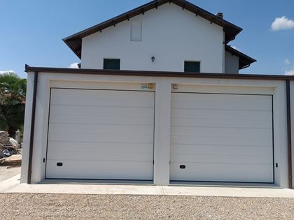 Cancello e recinzione in metallo bianco chiusi all'ingresso di un'abitazione, con pilastri in mattoni e vialetto pavimentato, sotto il sole.