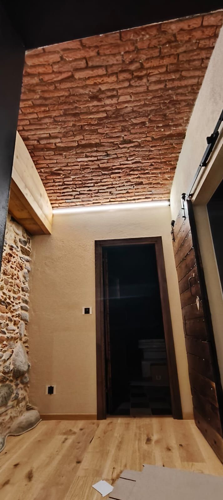 Stretto corridoio con soffitto in mattoni, pareti beige, pavimento in legno e una porta scura in fondo.
