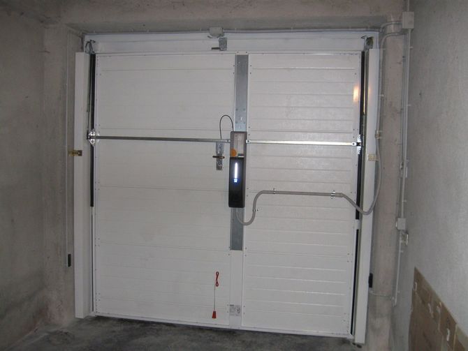 Doppie porte bianche in un corridoio di cemento, protette da una barra di chiusura verticale centrale.
