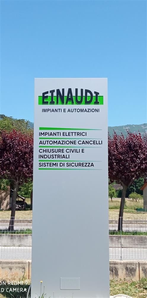 Cartello esterno per la scuola Einaudi, con testo in nero e verde su un pilastro grigio, su sfondo di cielo azzurro.