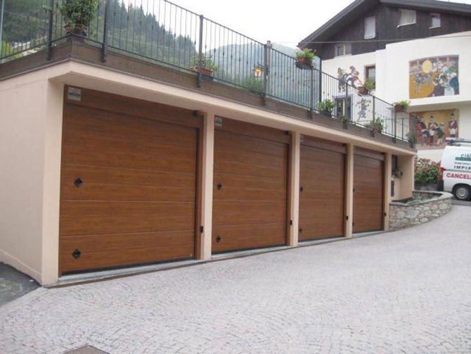 Una fila di tre porte da garage in legno lungo un edificio beige su un vialetto in pendenza lastricato.
