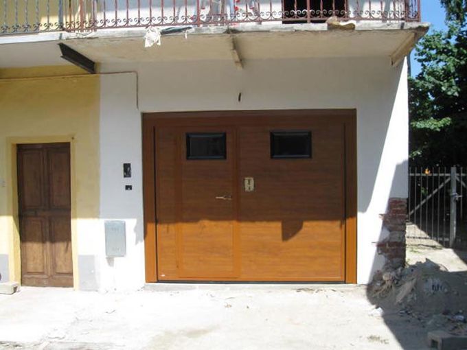 Facciata bianca della casa con un doppio portone da garage in legno e una piccola porta laterale all'ingresso.