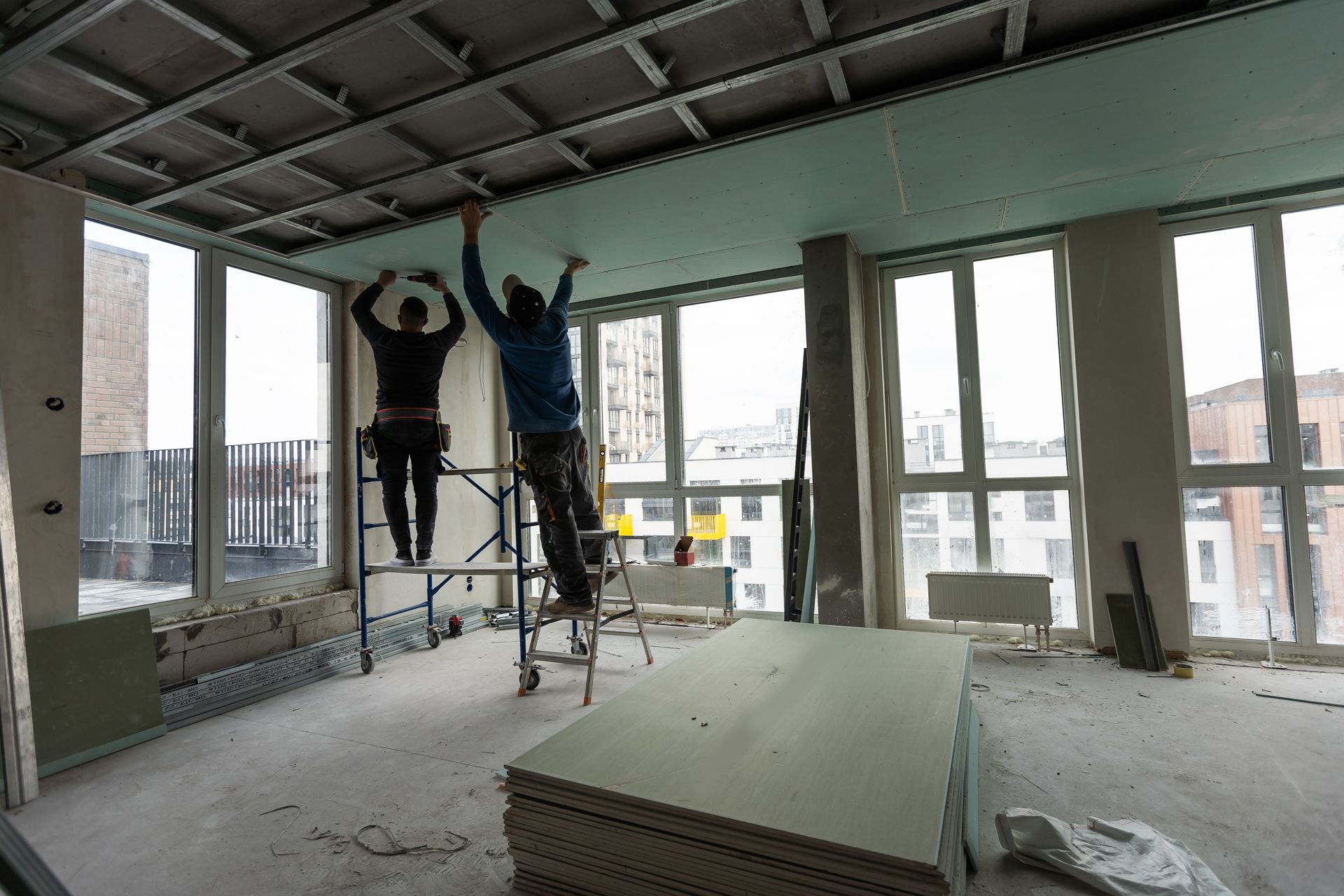 Two workers on scaffolding install green drywall panels onto a metal ceiling frame in a room with large windows.
