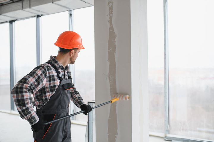A construction worker in a hard hat and work clothes uses a paint roller to paint a white pillar in a room with windows.