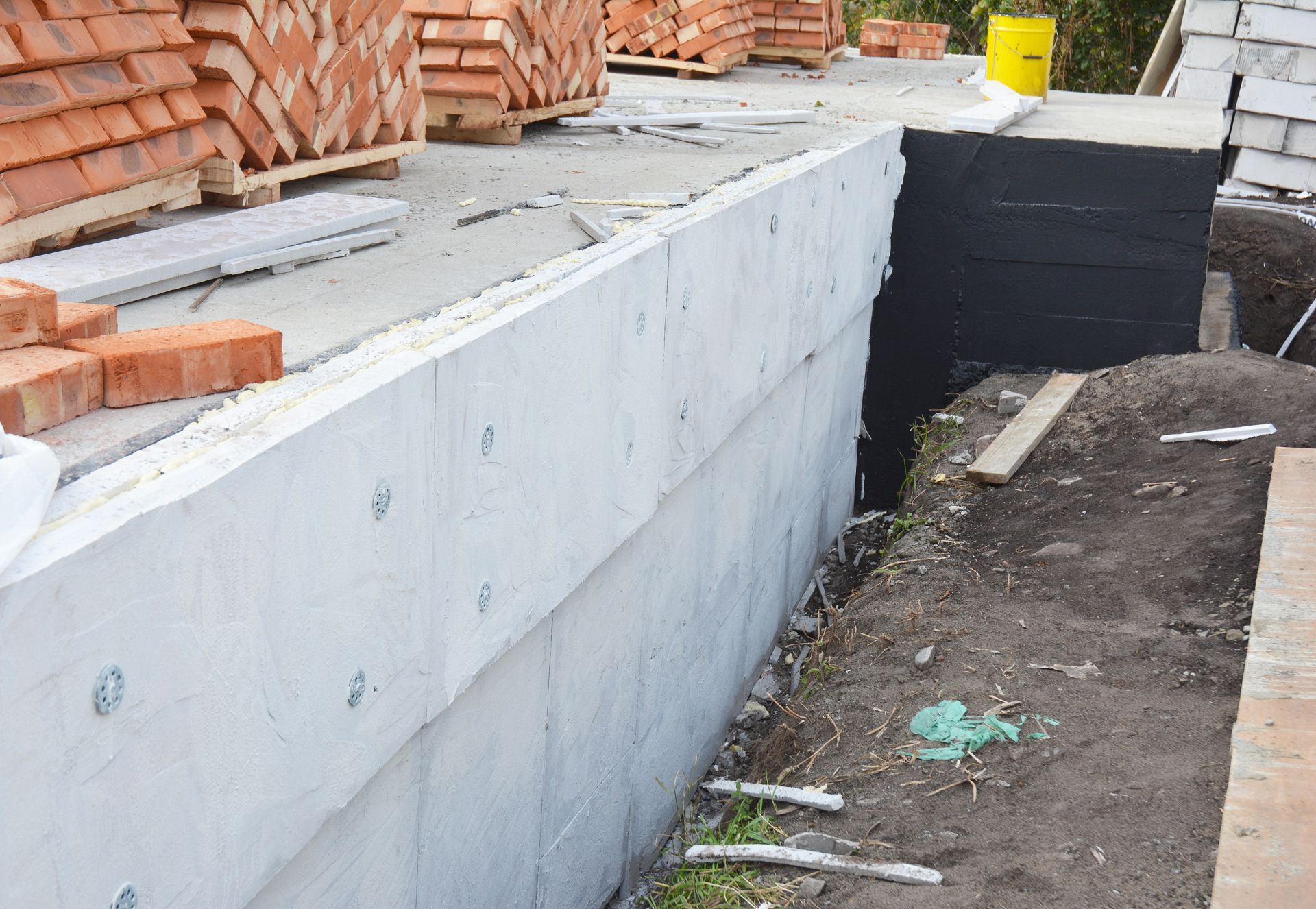 A concrete foundation wall stands next to a dirt trench, with stacks of red bricks on the flat surface above.