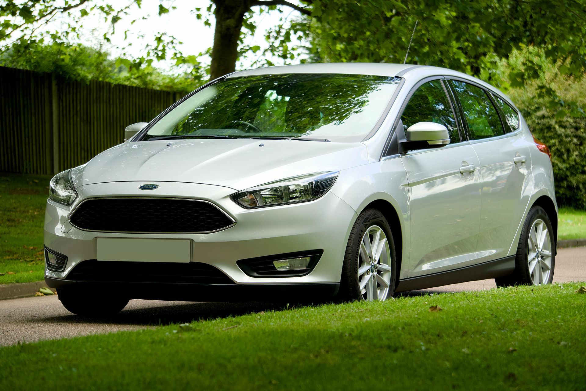 Silver hatchback parked on green grass beside a wooden fence in a leafy yard