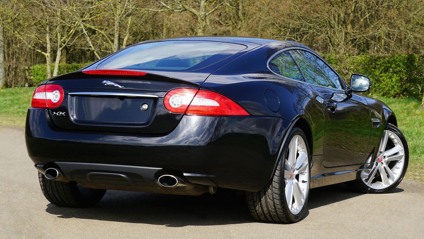 Black sports coupe parked outdoors on a paved road, rear-left view, with trees in the background