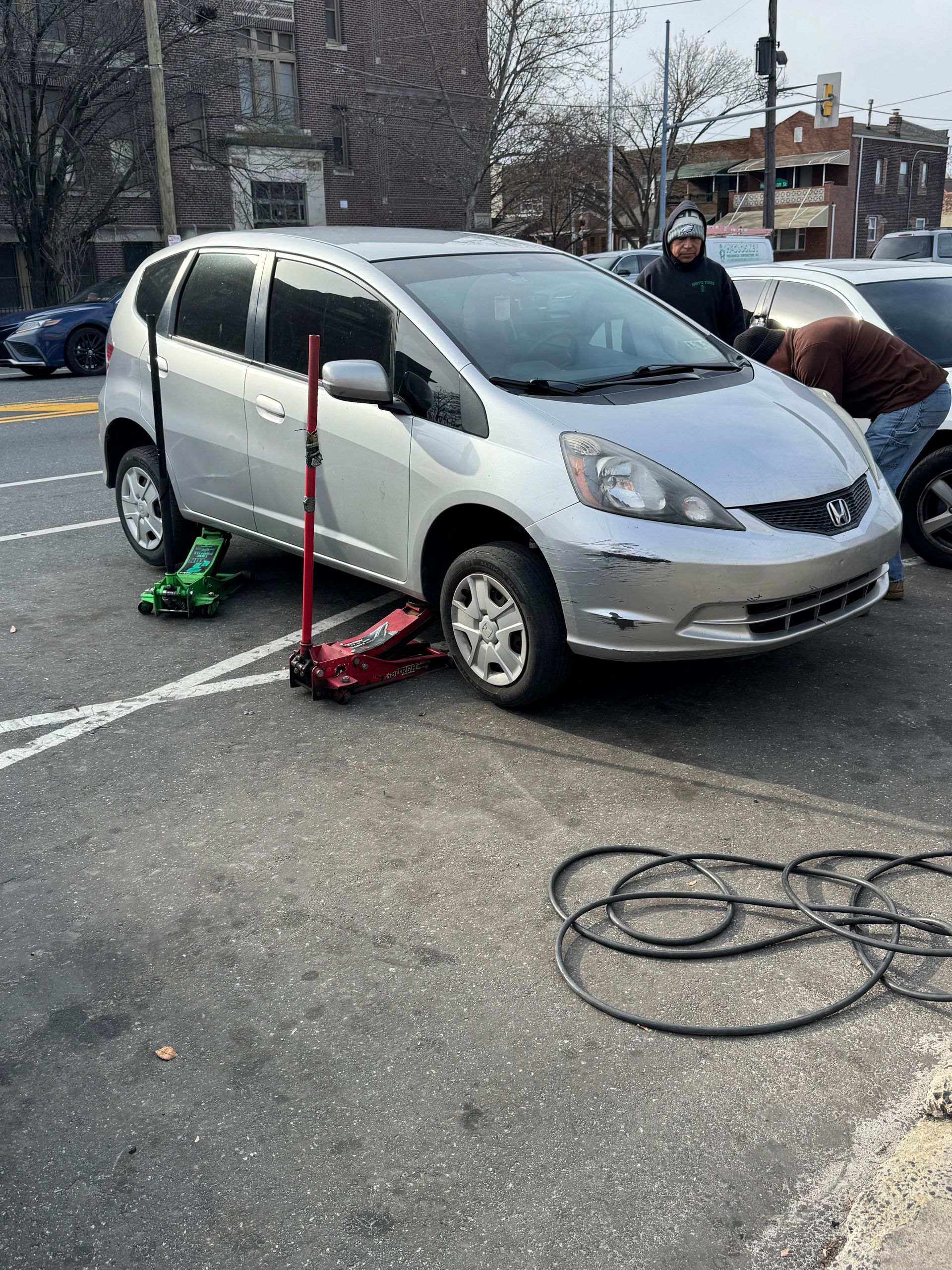 Silver minivan with a flat tire on a city street, workers using a jack and tools nearby.