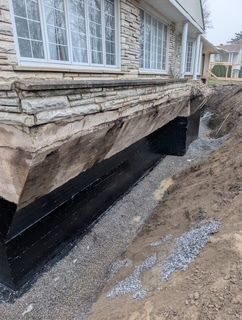 Fondations de maison présentant une fissure importante dans un mur et une membrane d'étanchéité noire apparente, à proximité d'une tranchée de fouille.