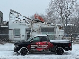 Une camionnette noire avec un texte publicitaire rouge est garée devant une maison enneigée endommagée et une clôture en grillage.