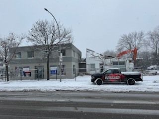 Rue enneigée avec une camionnette noire garée à côté d'un bâtiment gris et d'arbres dénudés.