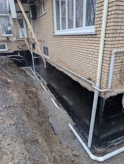 Étanchéité des fondations d'un bâtiment en briques avec tranchée apparente et tuyaux de drainage blancs près des fenêtres
