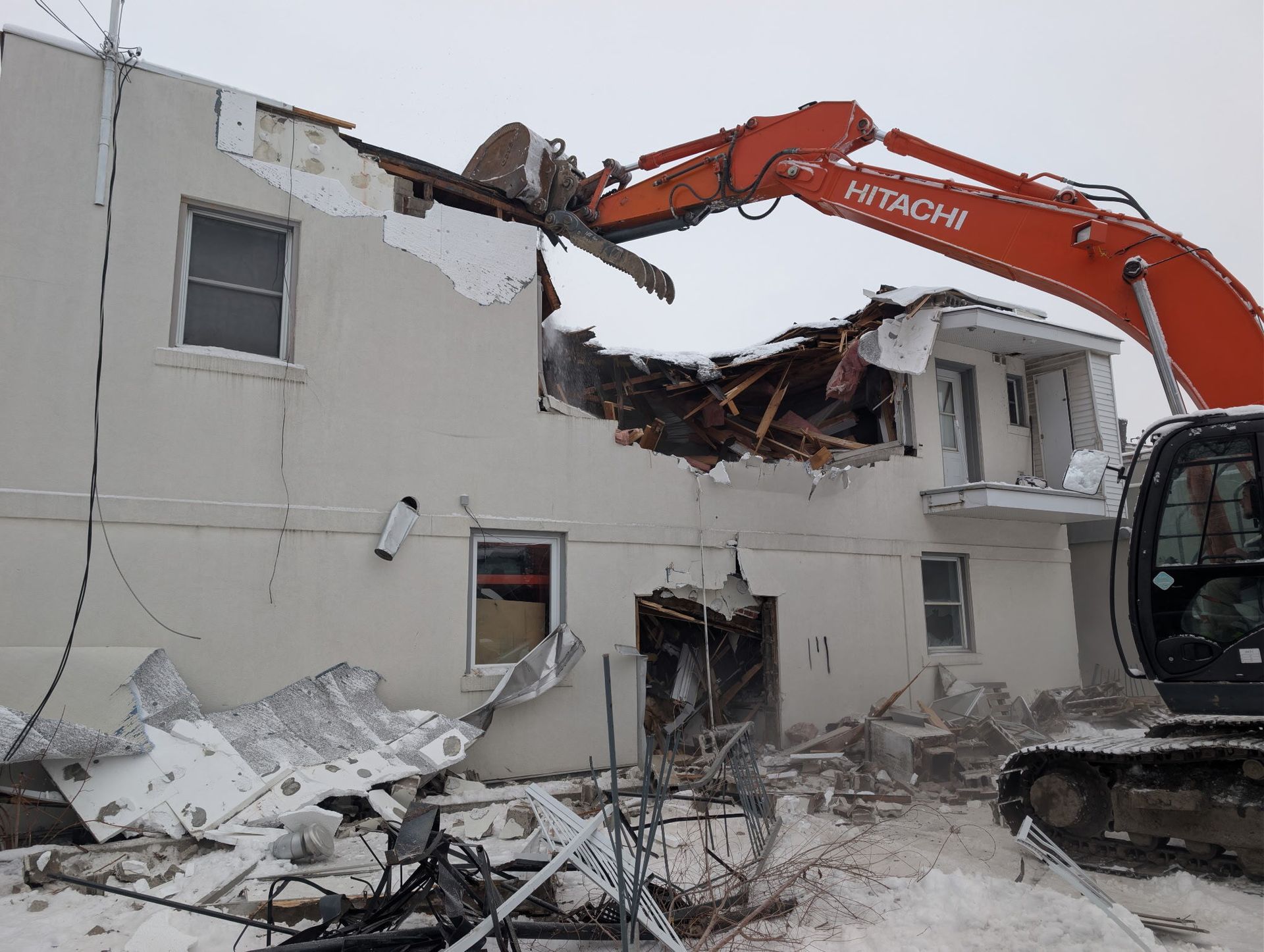 Une pelleteuse démolit un bâtiment blanc endommagé, laissant des décombres éparpillés sur le site.