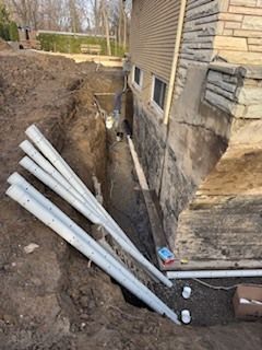 Tranchée de construction à côté de la maison, fondations apparentes et plusieurs tuyaux en PVC blanc gisant sur la pente de terre.