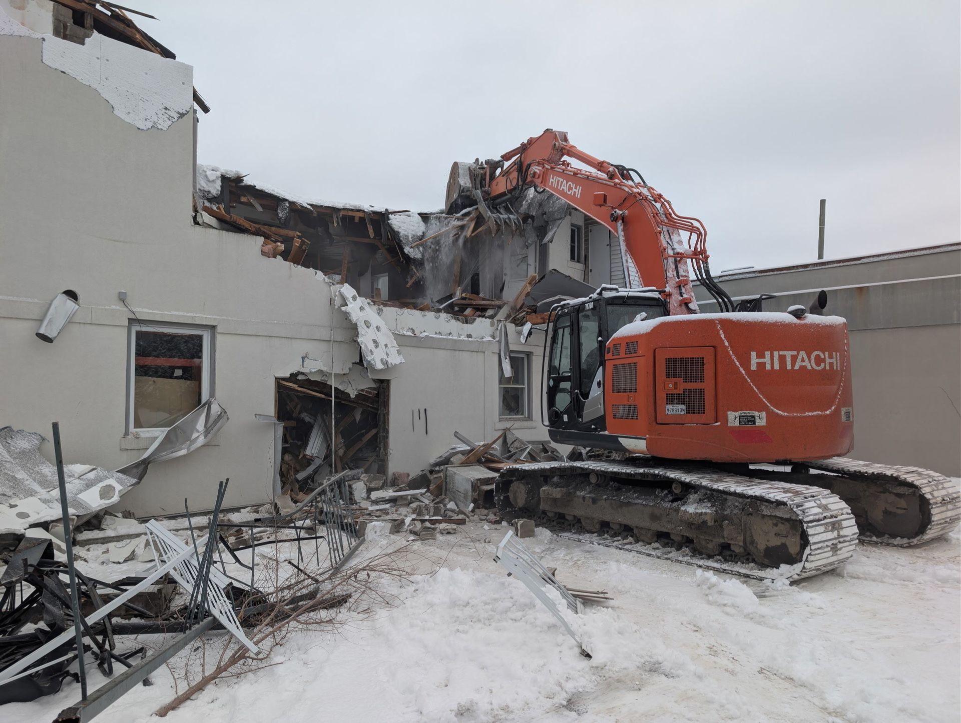 Une pelleteuse orange démolit un bâtiment blanc sur un site enneigé jonché de décombres