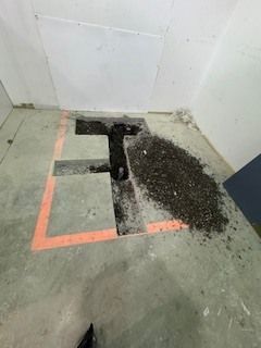 Ouverture marquée dans le sol avec une découpe noire en forme de T et un tas de débris dans une pièce d'angle blanche
