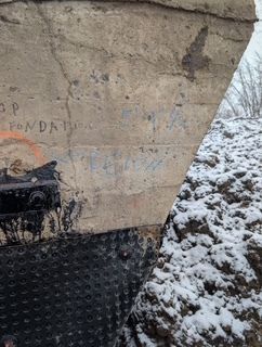 Pilier de pont en béton recouvert de neige, orné de graffitis et présentant une base texturée noire, à côté d'une pente enneigée.
