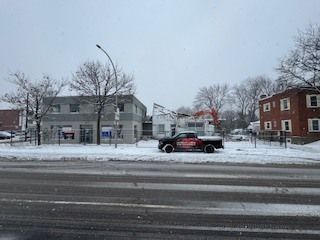 Rue enneigée avec une voiture noire passant devant des maisons grises et un bâtiment rouge par temps couvert