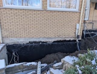 Excavation d'un mur de sous-sol à côté d'une maison en briques, mettant au jour les fondations et l'étanchéité noire.