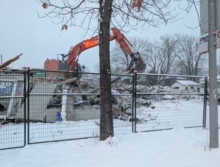 Une pelleteuse orange travaille derrière une clôture en grillage enneigée, près d'un arbre et d'un poteau électrique.
