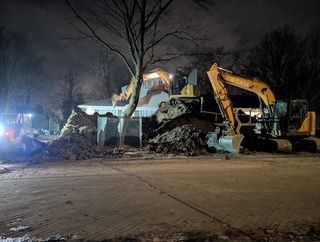 Des excavatrices jaunes dégagent de nuit un gros tas de terre en bordure de route enneigée.