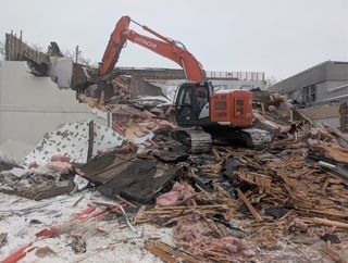 Une pelleteuse orange déblaye les gravats et les débris sur un chantier de démolition sous un ciel couvert.