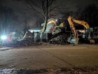Des excavatrices dégagent un tas de débris de nuit à côté d'une maison, la scène étant éclairée par de puissants projecteurs de travail.