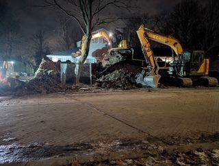 Scène de chantier nocturne avec des excavatrices déplaçant de la terre près d'un bâtiment éclairé et d'arbres dénudés.