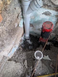 Zone de réparation de plomberie avec des tuyaux blancs apparents, un outil rouge et une bonde de sol dans un coin en béton brut